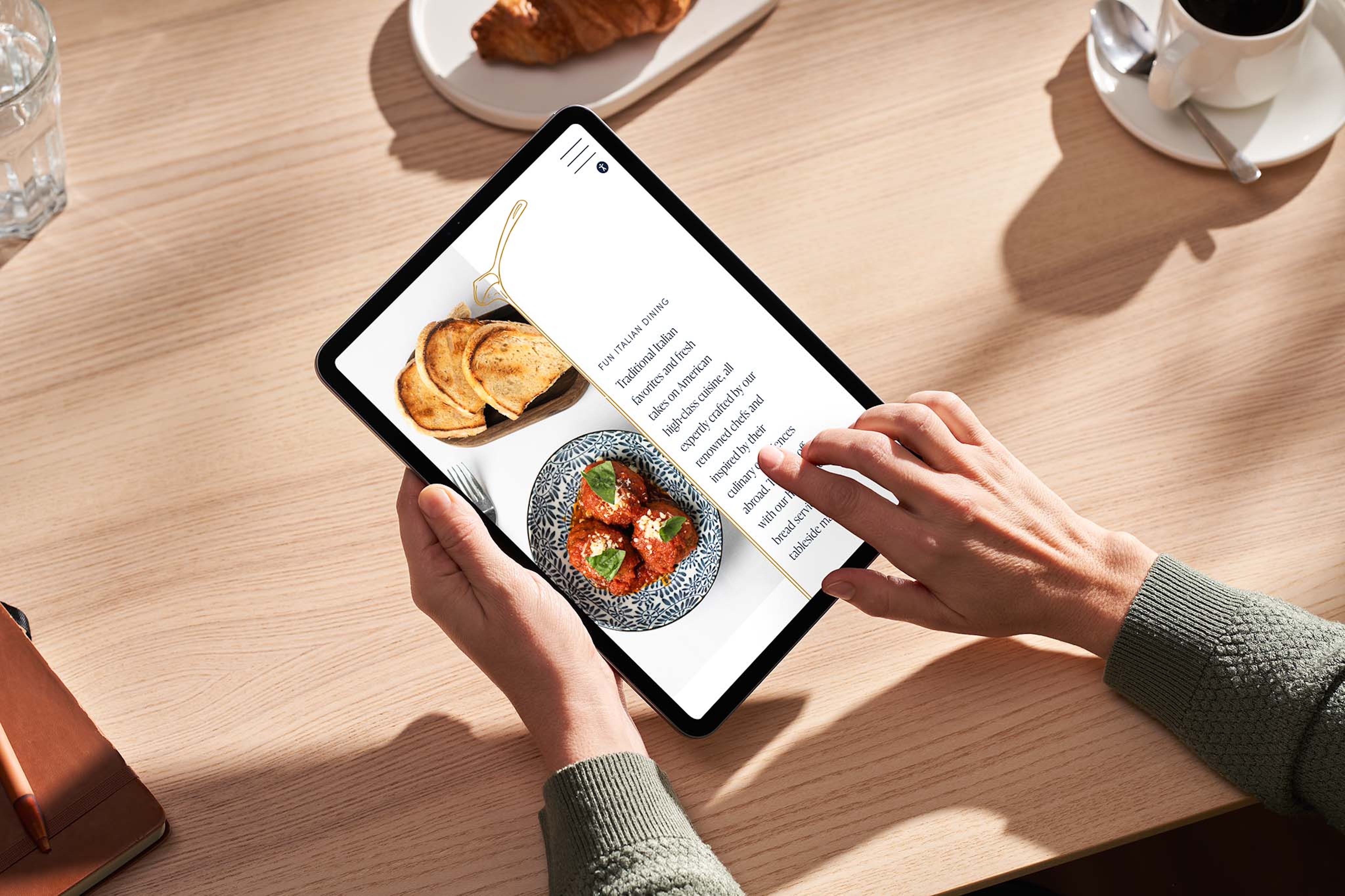 A person uses a tablet displaying a recipe with a photo of toast and a dish, sitting at a wooden table with coffee, a croissant, and water. The tablet's screen also shows the site's Privacy Policy link at the bottom.