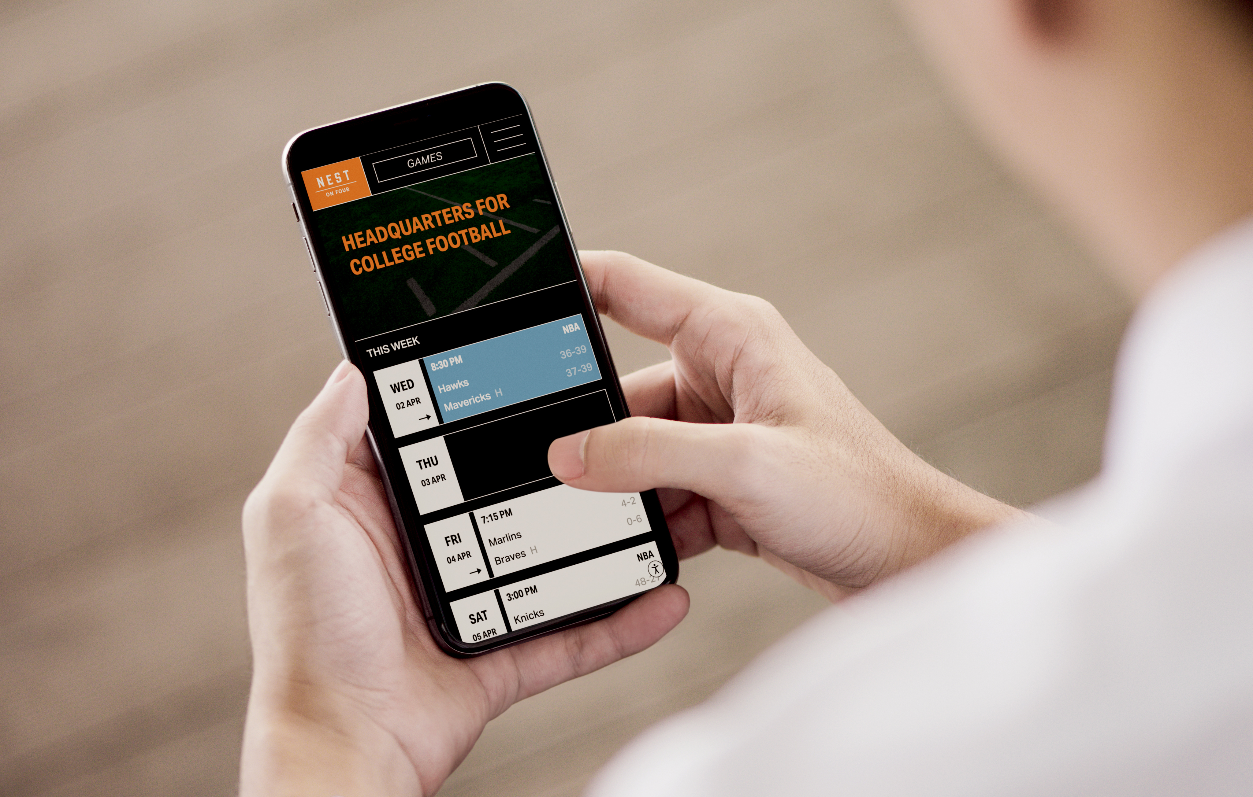 A person holding a smartphone displaying a turnkey college football schedule and results on a sports website or app.