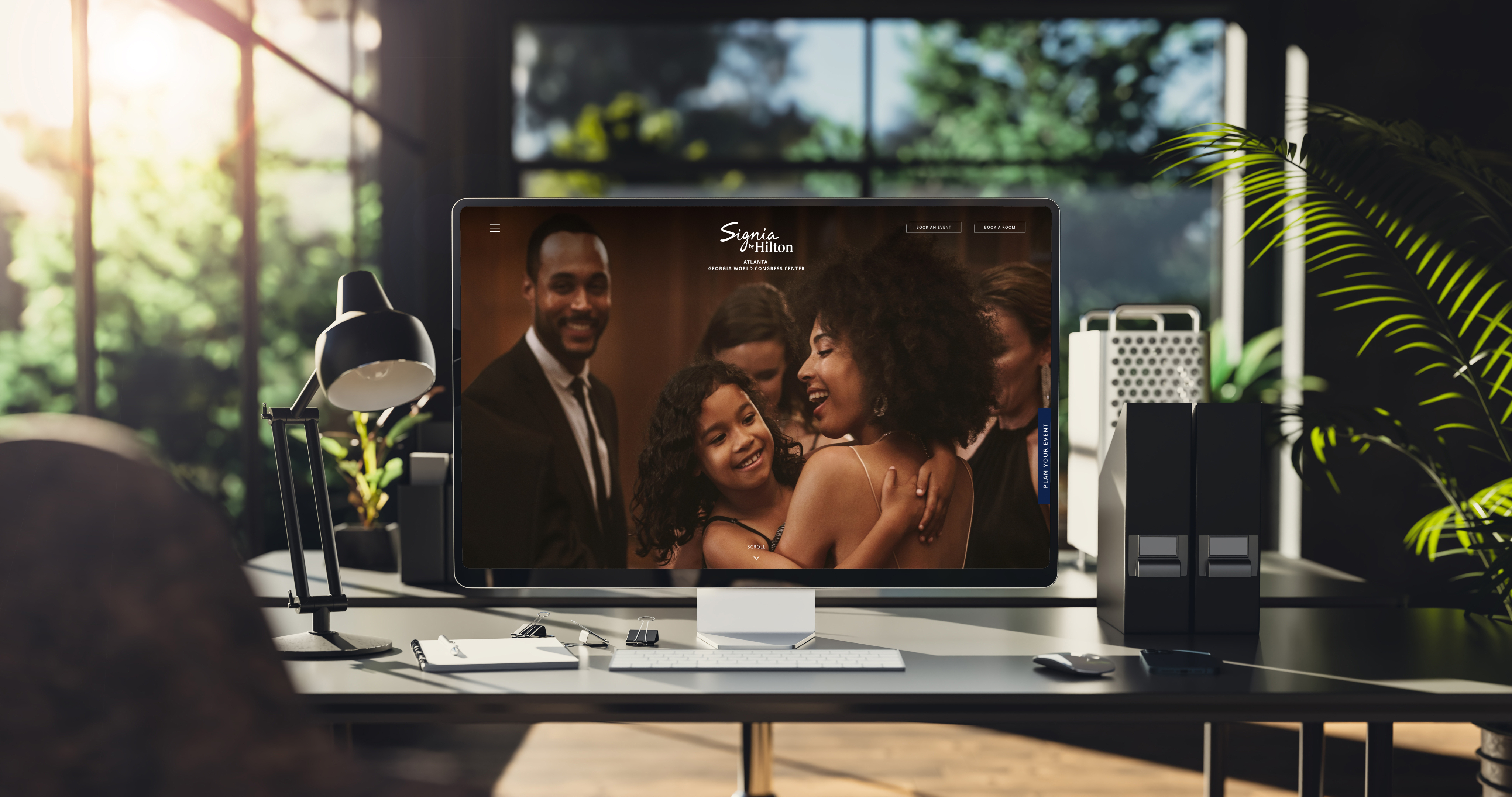 A modern turnkey office desk with a computer displaying a website featuring a smiling woman and child embracing at a social event. Sunlight streams through large windows in the background.