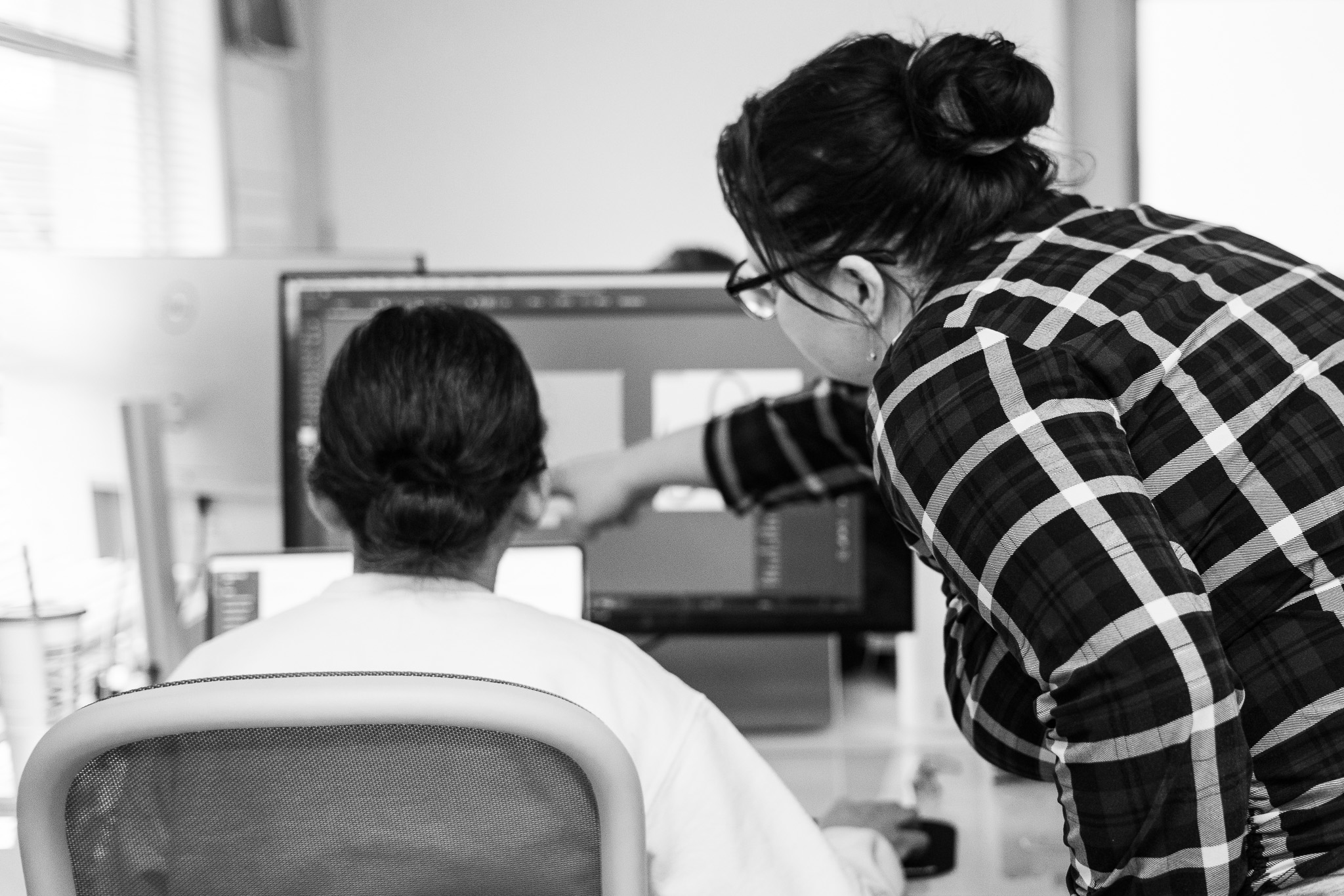 Two women work at computer monitors; one is seated while the other stands beside her, pointing at the screen.