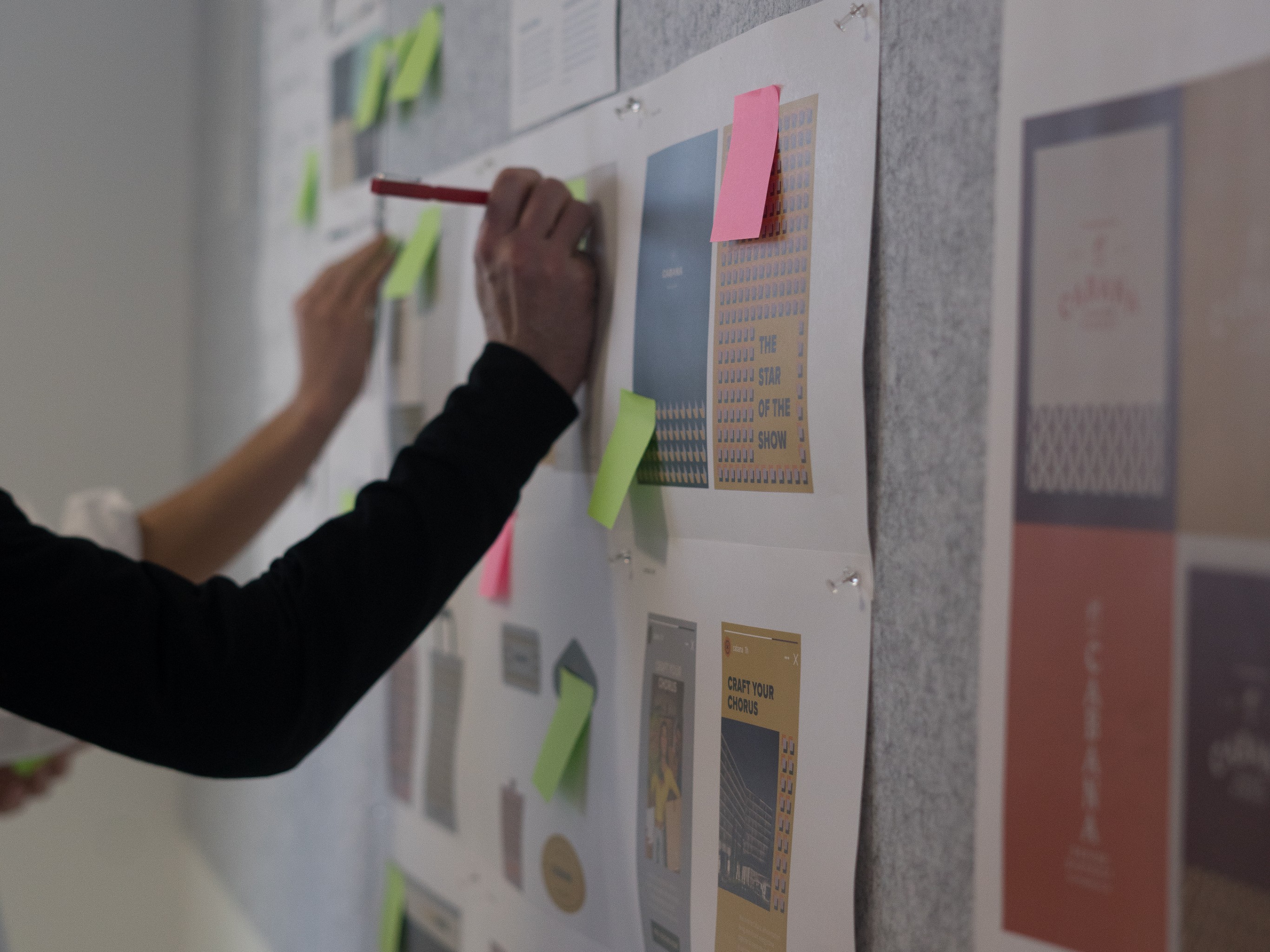 Two people place sticky notes and mark on printed pages with images and text pinned to a wall, suggesting a planning or brainstorming session.